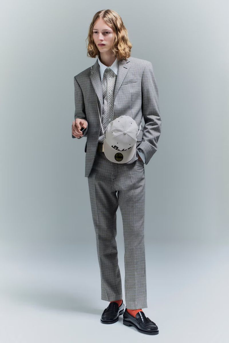 Full-length shot of a young adult male fashion model in a houndstooth suit, standing against a gray background with a baseball cap bag.
