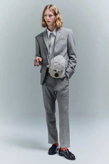Full-length shot of a young adult male fashion model in a houndstooth suit, standing against a gray background with a baseball cap bag.