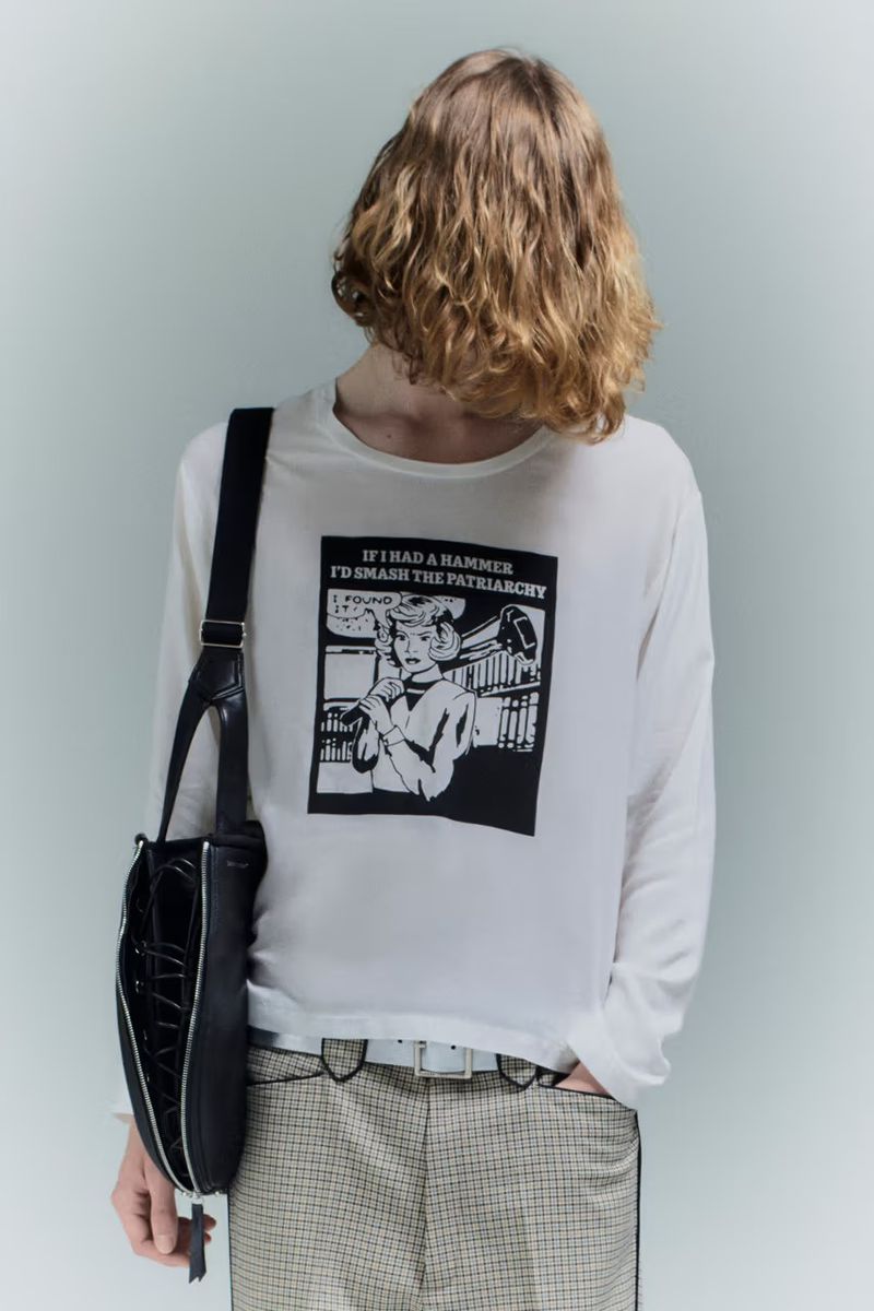 Back view of an adult wearing a white long-sleeve shirt featuring black graphic art and a black purse slung over the shoulder.