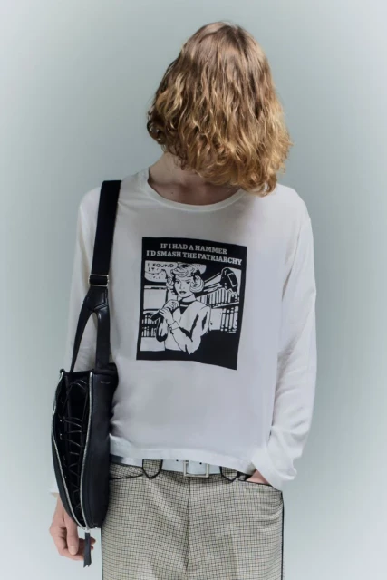 Back view of an adult wearing a white long-sleeve shirt featuring black graphic art and a black purse slung over the shoulder.