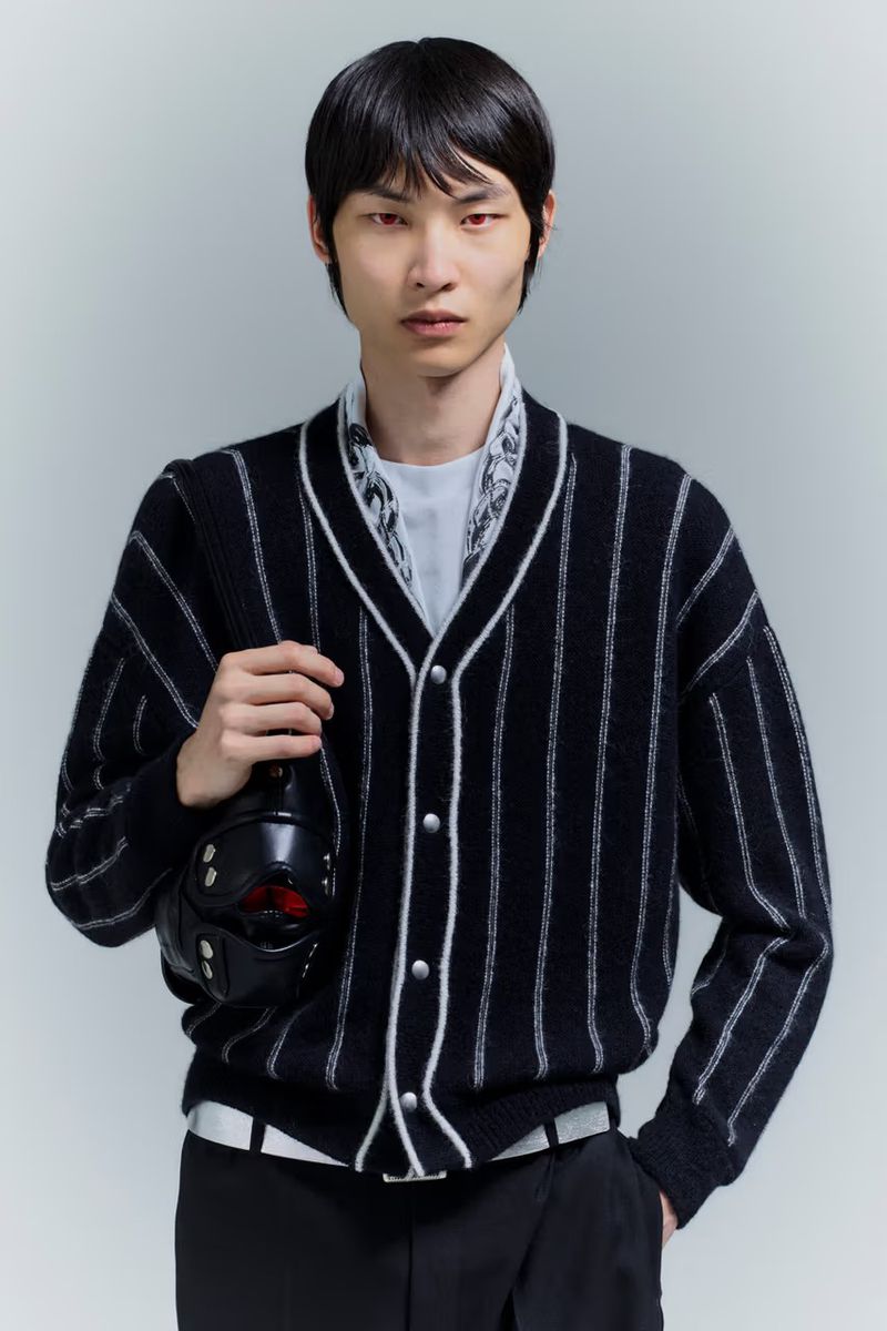 Asian male model poses indoors wearing a vertically striped cardigan, dark pants, and carrying a black helmet-like object in hand.