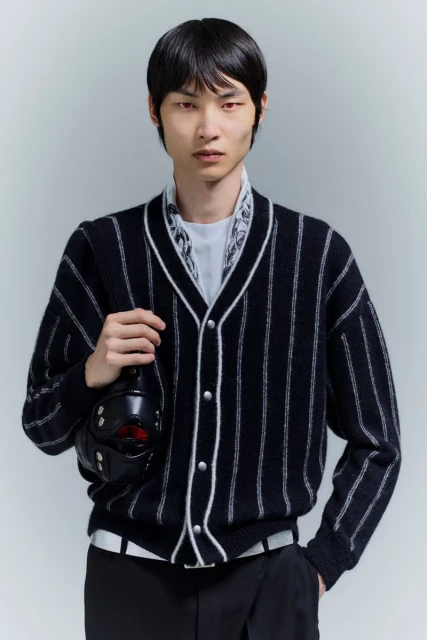 Asian male model poses indoors wearing a vertically striped cardigan, dark pants, and carrying a black helmet-like object in hand.