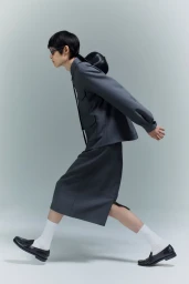 Model in grey shirt, skirt, white socks and black loafers striding against a grey studio seamless background.