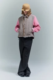 Model wearing a patterned jacket with pink sleeves, dark trousers, and a tan cap against a grey studio backdrop.