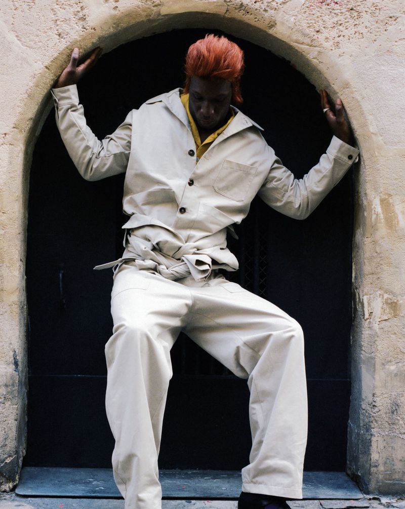 Stylish adult male poses confidently in an open doorway wearing a sand-colored jacket and baggy pants, showcasing a fashion-forward look.