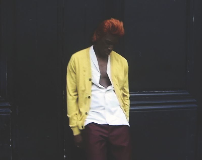 Young man with orange hair poses against a dark wall wearing a yellow cardigan, white shirt, and maroon pants in an urban setting.