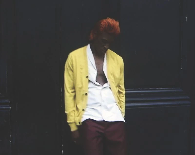 Young man with orange hair poses against a dark wall wearing a yellow cardigan, white shirt, and maroon pants in an urban setting.