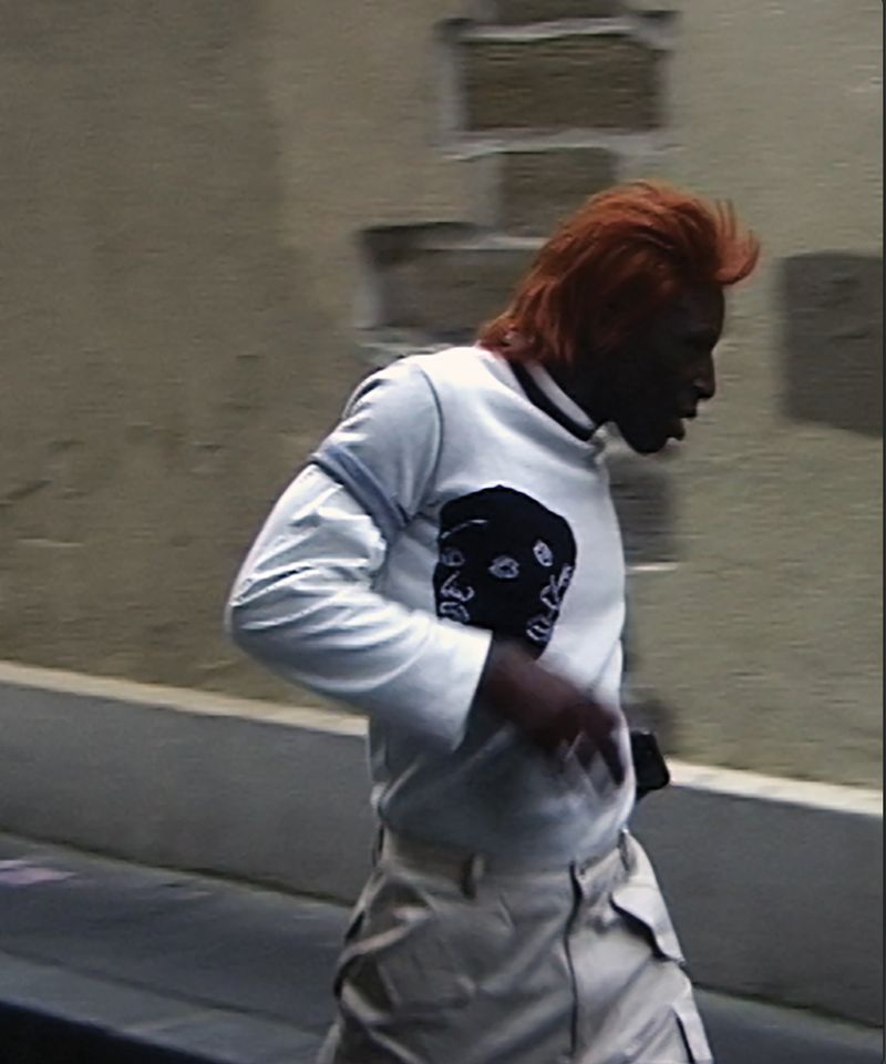 Adult male walking near a building with orange hair, wearing a white long sleeve shirt with a graphic and light colored pants.