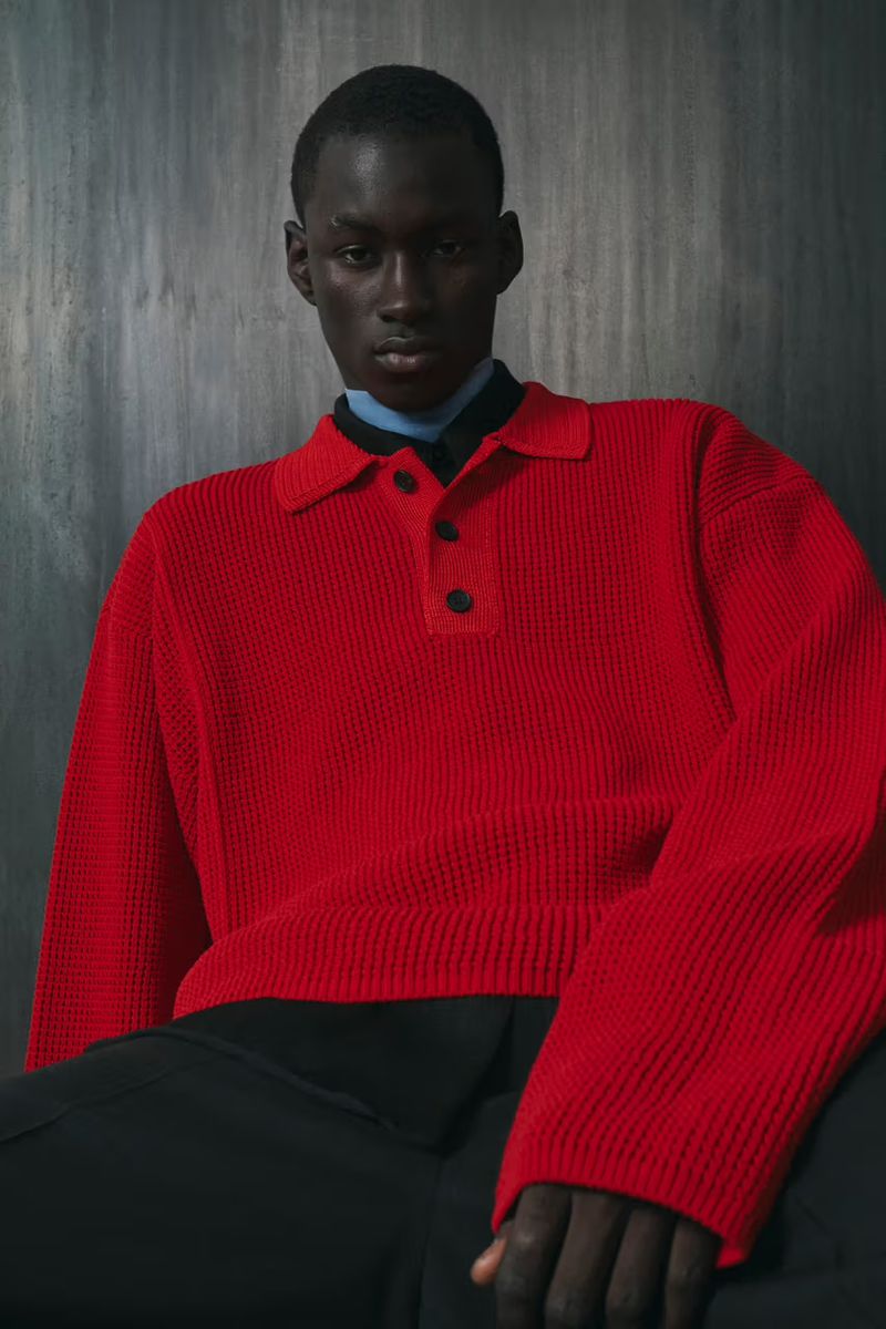 Portrait of a dark-skinned adult male wearing a red knit collared shirt against a grey textured background, looking at the camera.