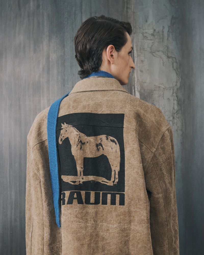 Adult wearing a tan jacket with a horse graphic on the back poses against a grey wall indoors in a fashion editorial shot.