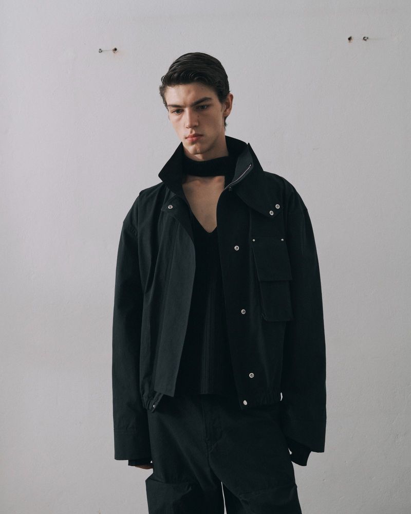 A light-skinned adult male models a black jacket, v-neck sweater, and pants against a plain white wall in a studio setting.