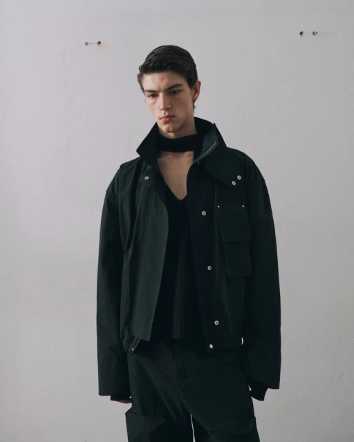 A light-skinned adult male models a black jacket, v-neck sweater, and pants against a plain white wall in a studio setting.