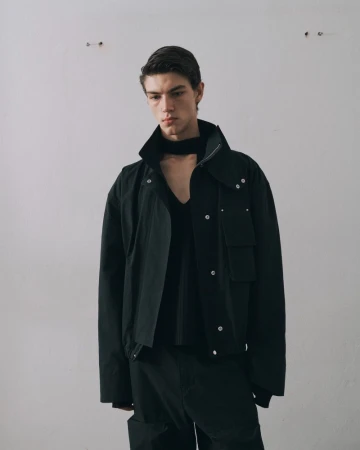 A light-skinned adult male models a black jacket, v-neck sweater, and pants against a plain white wall in a studio setting.