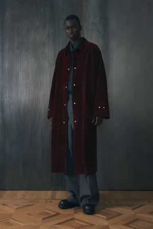 Full-length shot of an African-American man in a maroon overcoat, gray trousers and black shoes, posing against a textured gray backdrop.