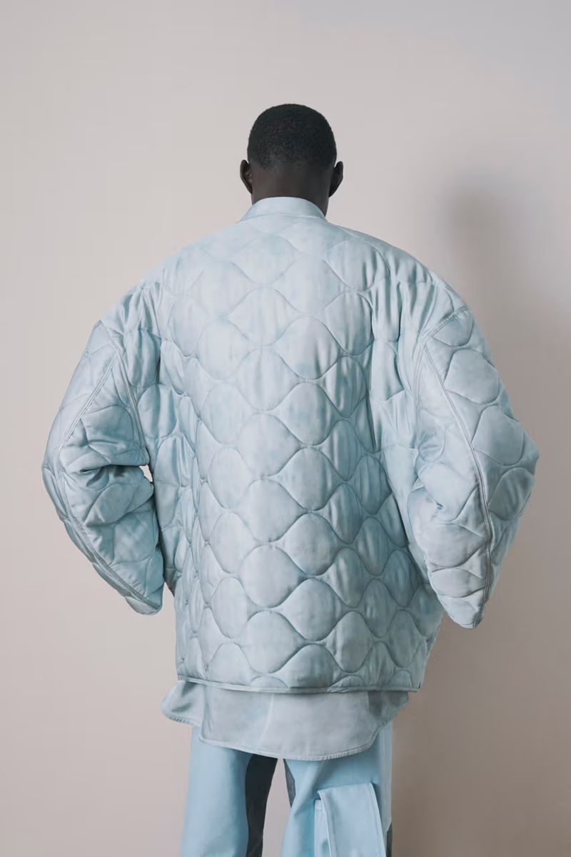Back view of a dark-skinned man wearing a quilted light blue jacket and light blue trousers, standing against an off-white background.
