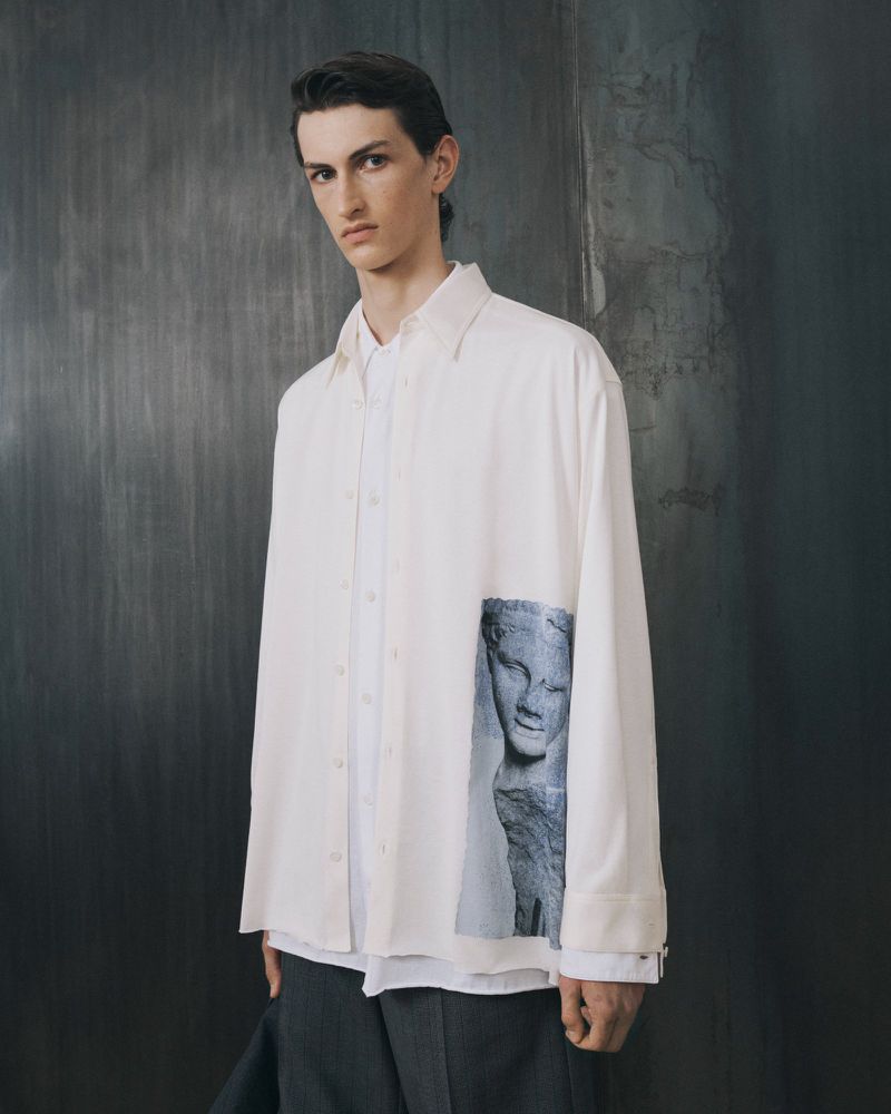 A young adult male poses in a white button-down shirt with a graphic print, paired with grey slacks, against a textured backdrop.