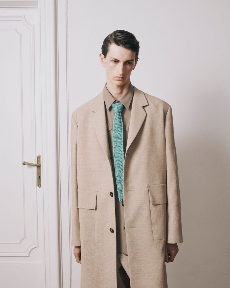 Young adult male fashion model in a beige trench coat and a teal knit tie poses in front of a white door for a studio fashion shoot.