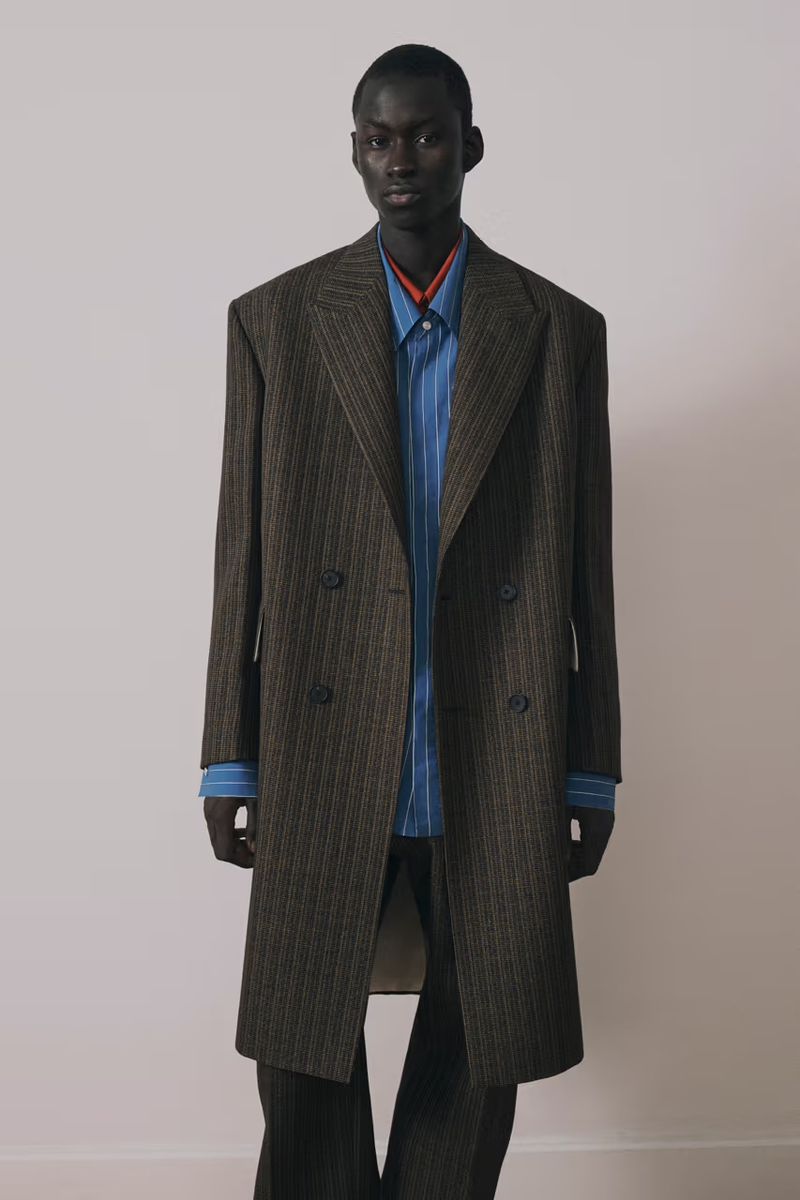 A dark-skinned man in a striped suit stands against a white wall, wearing a blue striped shirt with a red beaded necklace. He is looking at the camera.