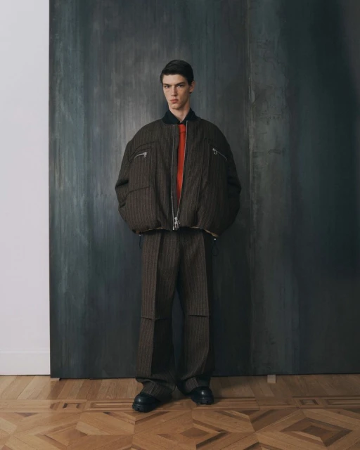 A young man stands in a brown pinstripe suit and bomber jacket in front of a textured gray background, with wood floors.
