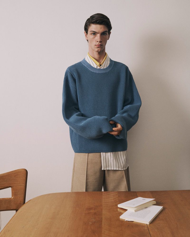 Portrait of a young male model in a blue sweater and beige trousers, standing in front of a neutral wall with a wooden table and books.