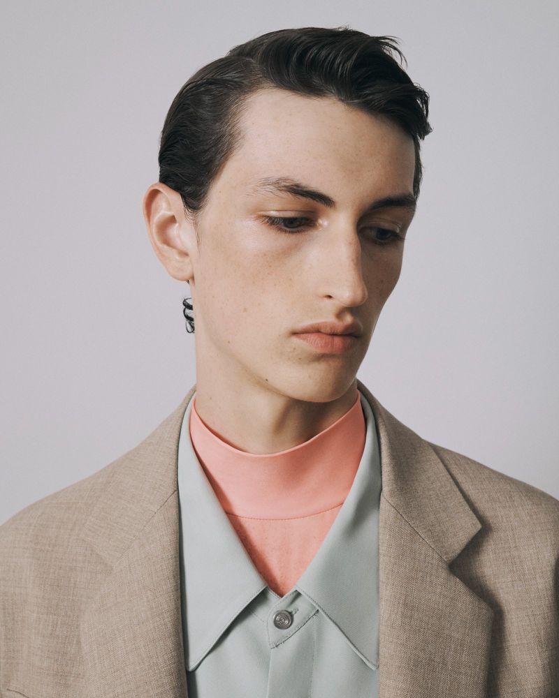 Portrait of a young adult male in a blazer with a colorful shirt and dark earring looking downward against a neutral background.