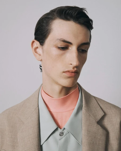 Portrait of a young adult male in a blazer with a colorful shirt and dark earring looking downward against a neutral background.