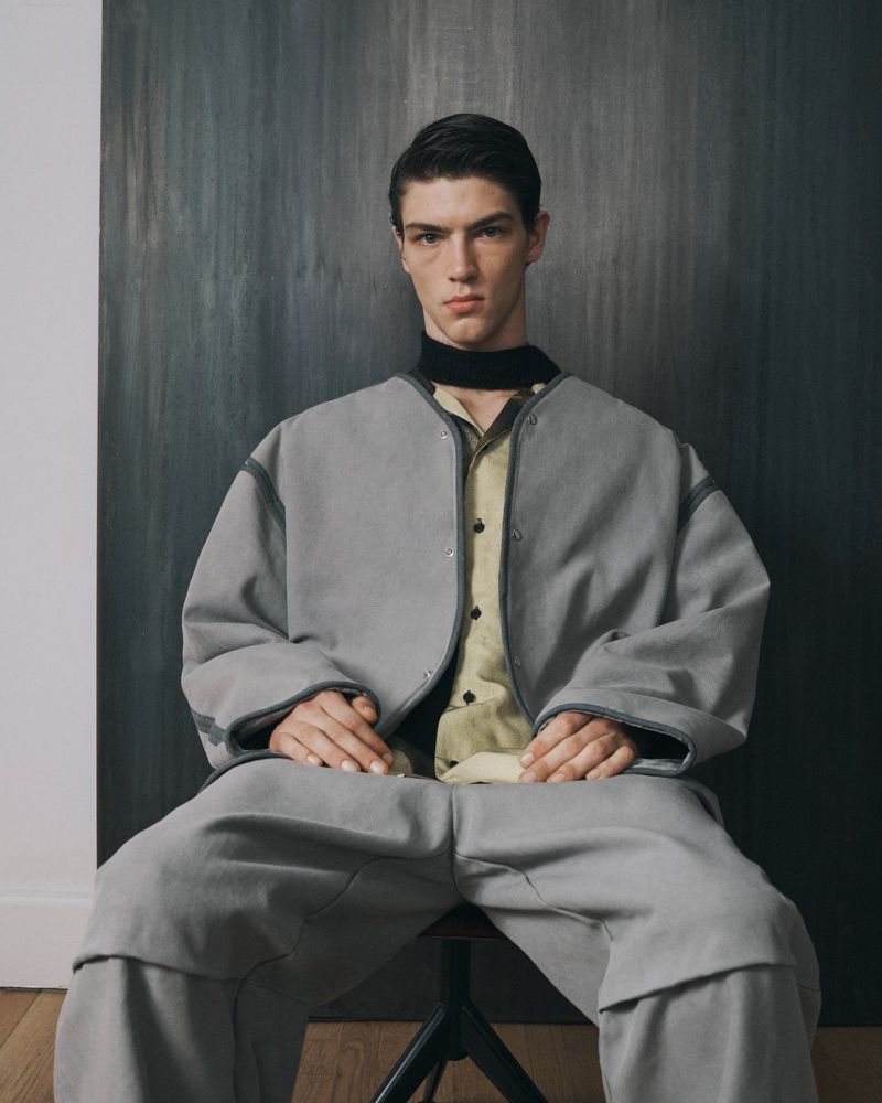 Portrait of a young man in a gray jacket and pants sitting on a black stool against a grey textured wall. Fashion editorial shot.