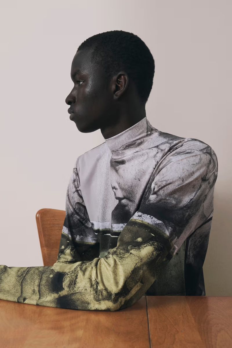 Profile of a dark-skinned man with short hair, wearing a printed shirt while seated at a table in an indoor setting against a light wall.