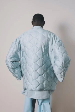 Model seen from behind wearing an oversized quilted pale blue jacket and matching trousers against a neutral studio backdrop.