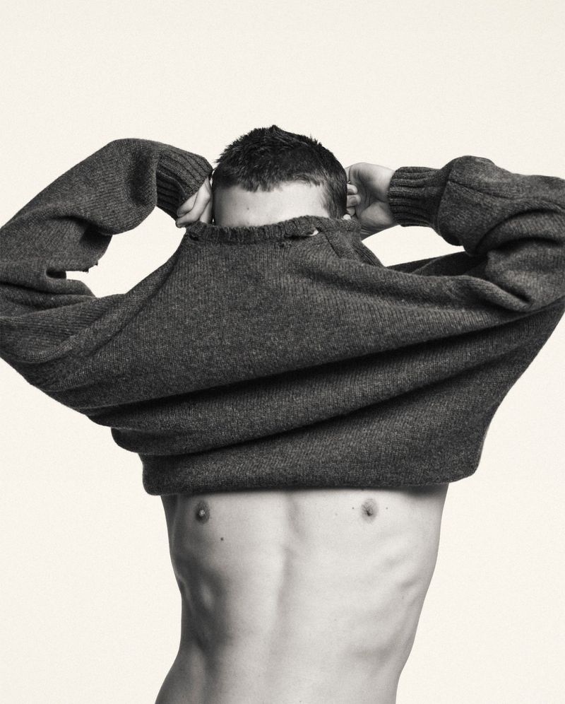 Monochrome portrait of a topless man with a dark sweater over his head, posing against an off-white background in a photo studio.