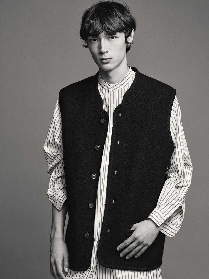 Portrait of a young adult male wearing a black vest over a striped shirt, posing in a studio with a neutral background.