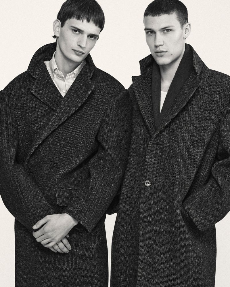 Two adult male models wearing matching dark grey wool overcoats, indoor setting with neutral backdrop. Fashion editorial shot.