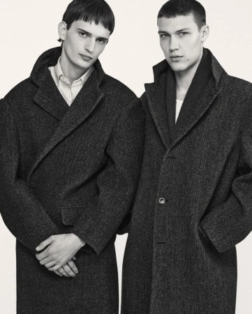 Two adult male models wearing matching dark grey wool overcoats, indoor setting with neutral backdrop. Fashion editorial shot.