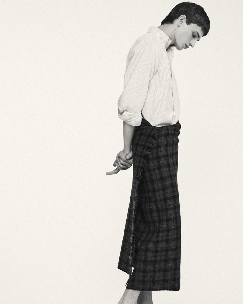 Monochrome studio shot: Man in white shirt and plaid wrap skirt standing with hands clasped behind, minimal aesthetic.