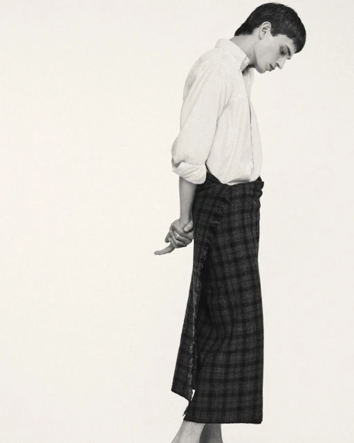 Monochrome studio shot: Man in white shirt and plaid wrap skirt standing with hands clasped behind, minimal aesthetic.
