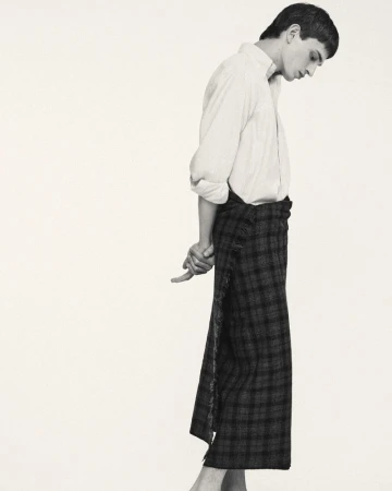 Monochrome studio shot: Man in white shirt and plaid wrap skirt standing with hands clasped behind, minimal aesthetic.