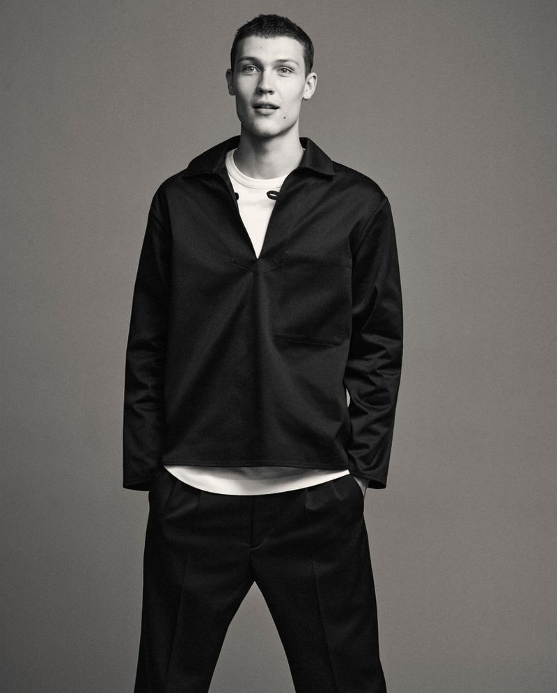 Male model in a black collared shirt and trousers standing against a plain grey studio backdrop