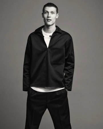 Male model in a black collared shirt and trousers standing against a plain grey studio backdrop