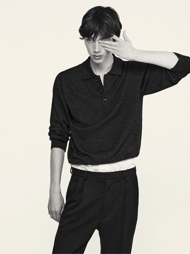 A young male model wearing a dark polo shirt and black trousers, lifting his hand to cover one eye.