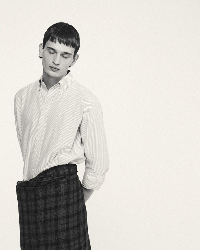Monochromatic portrait of a young adult man with short hair wearing a button-down shirt and a plaid skirt looking down indoors.