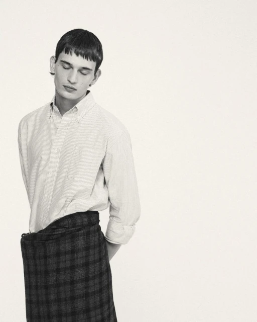 Monochromatic portrait of a young adult man with short hair wearing a button-down shirt and a plaid skirt looking down indoors.