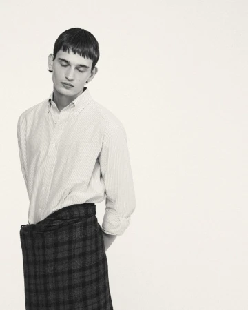 Monochromatic portrait of a young adult man with short hair wearing a button-down shirt and a plaid skirt looking down indoors.