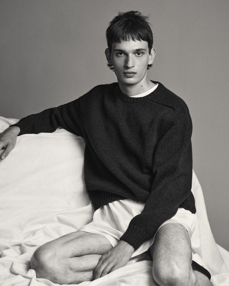 A young male model wearing a dark sweater and white shorts seated against a seamless grey backdrop.