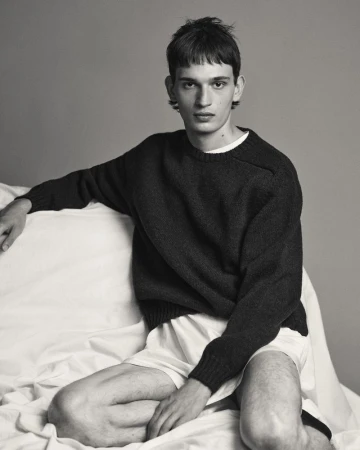 A young male model wearing a dark sweater and white shorts seated against a seamless grey backdrop.