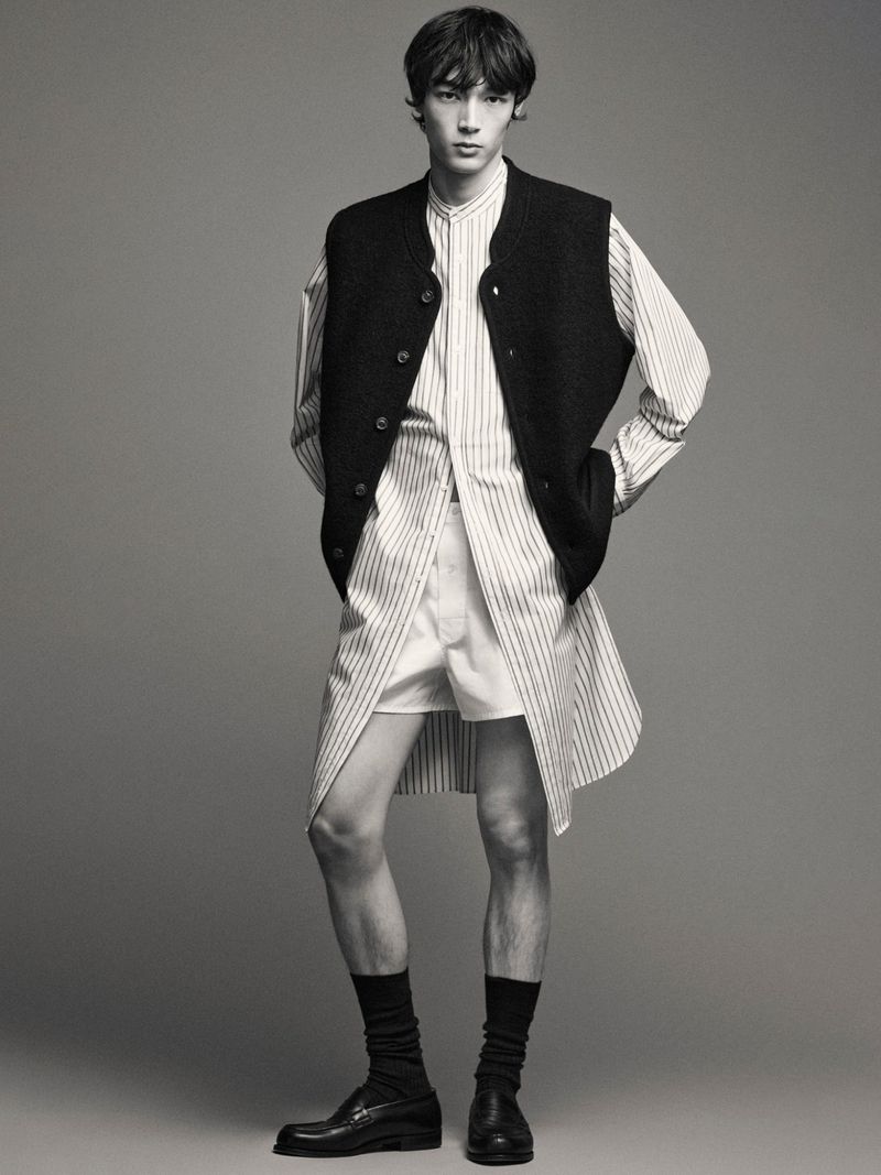 Male fashion model poses in a vertically striped shirt, vest, shorts, socks, and loafers against a gray studio backdrop.