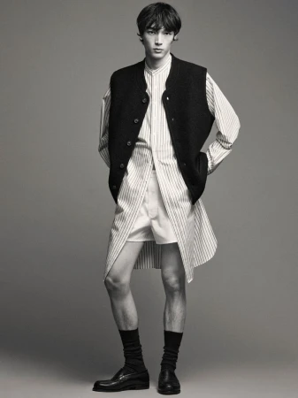 Male fashion model poses in a vertically striped shirt, vest, shorts, socks, and loafers against a gray studio backdrop.