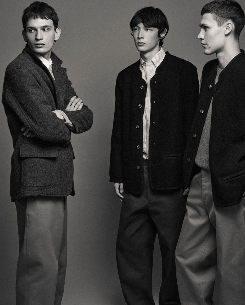 Three adult male models stand in a studio wearing dark jackets and trousers. A vertical fashion-style studio shot with a neutral grey background.