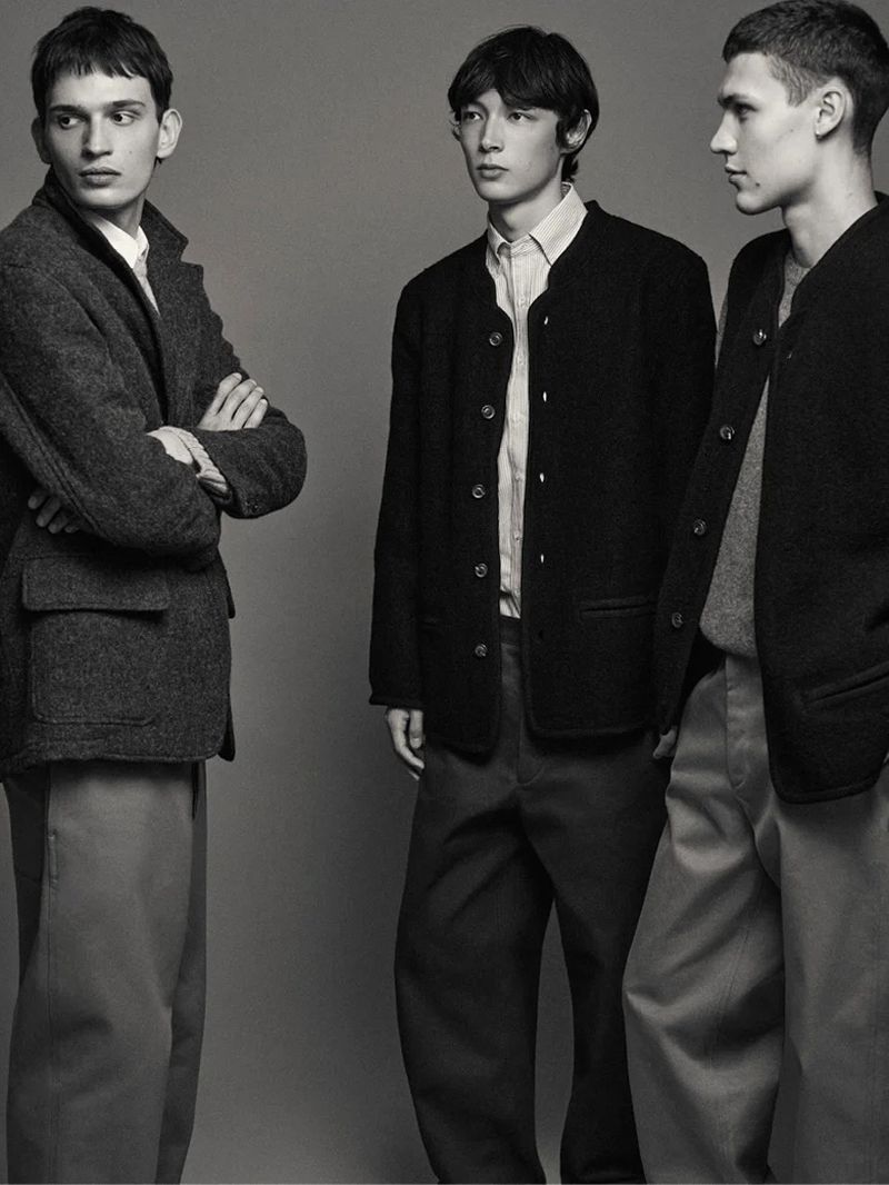 Three young men pose in a studio, showcasing wool coats and trousers in a monochromatic grey fashion editorial. 