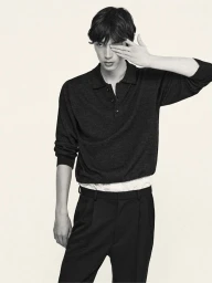 A young male model wearing a dark polo shirt and black trousers, lifting his hand to cover one eye.