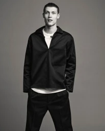 Male model in a black collared shirt and trousers standing against a plain grey studio backdrop